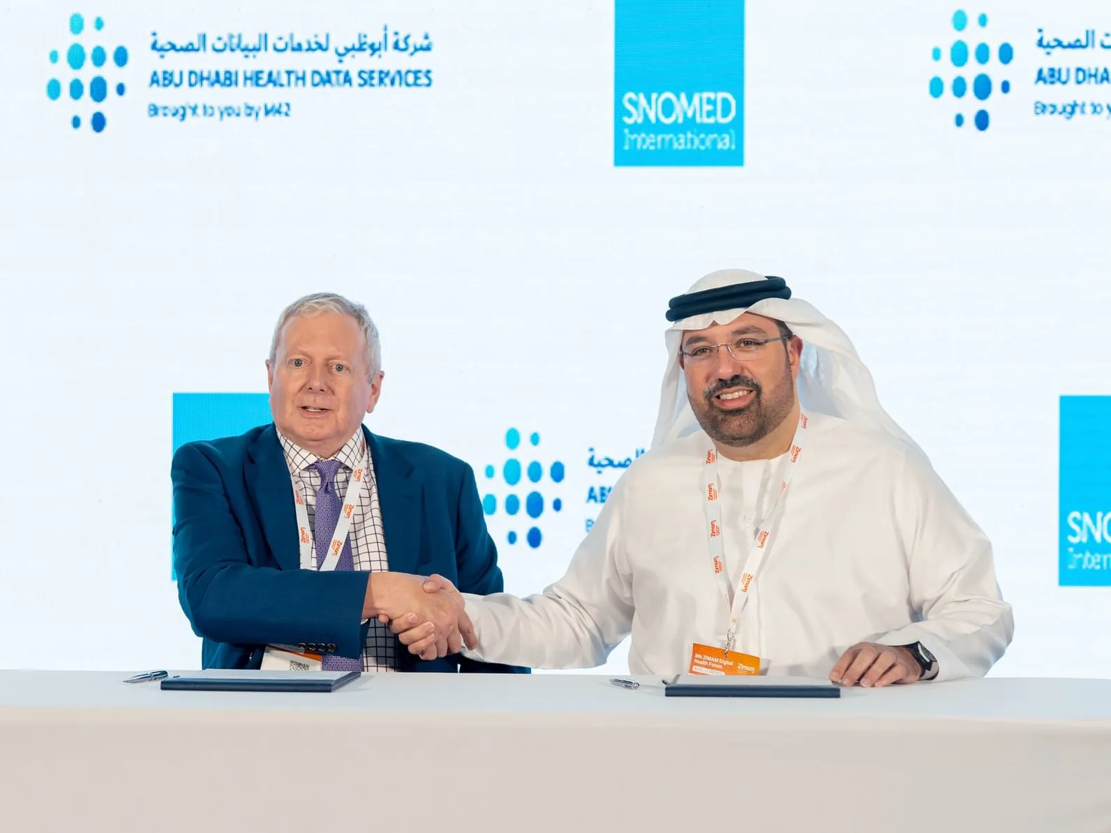 Two men, one in a suit and the other in traditional Arabic attire, shake hands in front of a backdrop with company logos.