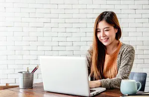 A smiling young Asian woman with long brown hair works on a silver laptop at a wooden desk with a white brick wall in the background.