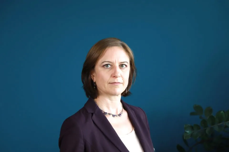 A woman with short brown hair, wearing a purple blazer and a white top, looks directly at the camera against a blue background with a plant in the lower right corner.