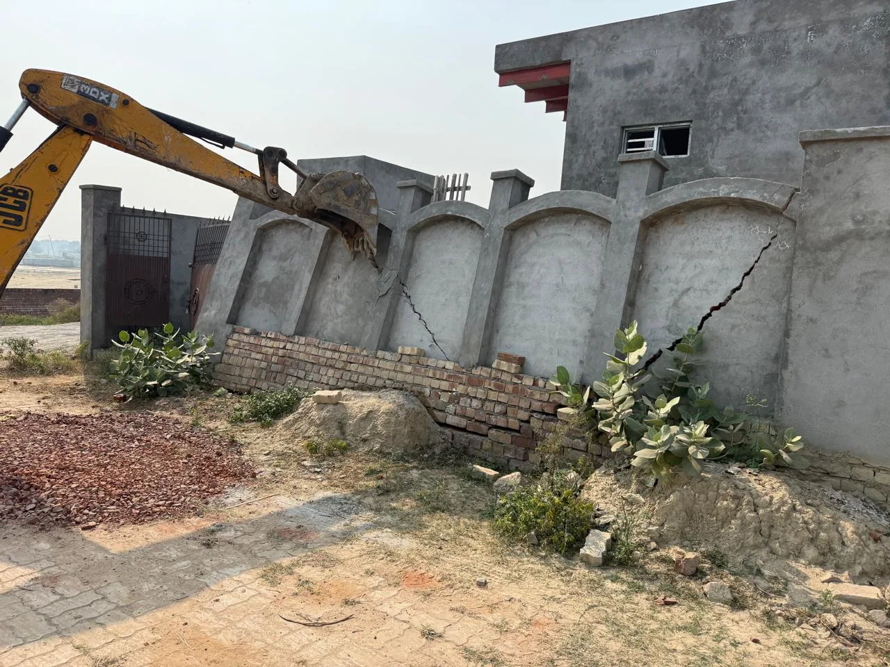 A yellow JCB excavator arm with a bucket positioned to demolish a gray concrete boundary wall showing large vertical cracks, adjacent to an under-construction or partially damaged building.