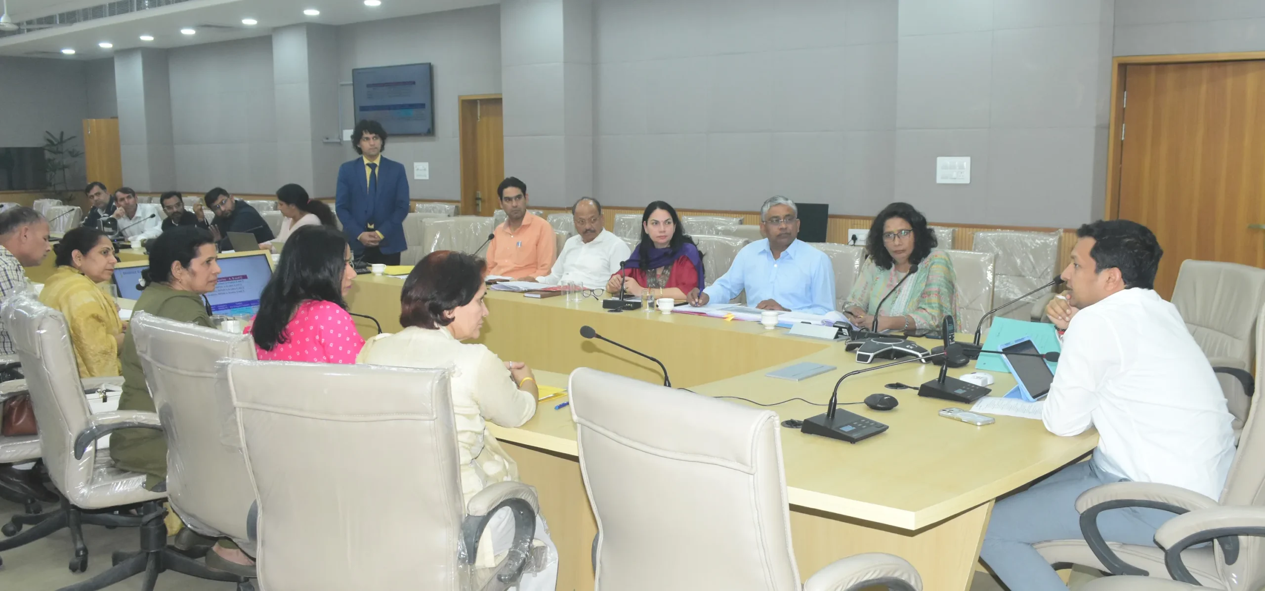 Deputy-Commissioner-Sachin-Gupta-announing-the-mission-during-a-high-level-review-meeting-held-at-the-Mini-Secretariat attend a formal high-level committee meeting in a conference room, seated around a large table with individual microphones. A presenter in a blue suit stands near a presentation screen on the left.