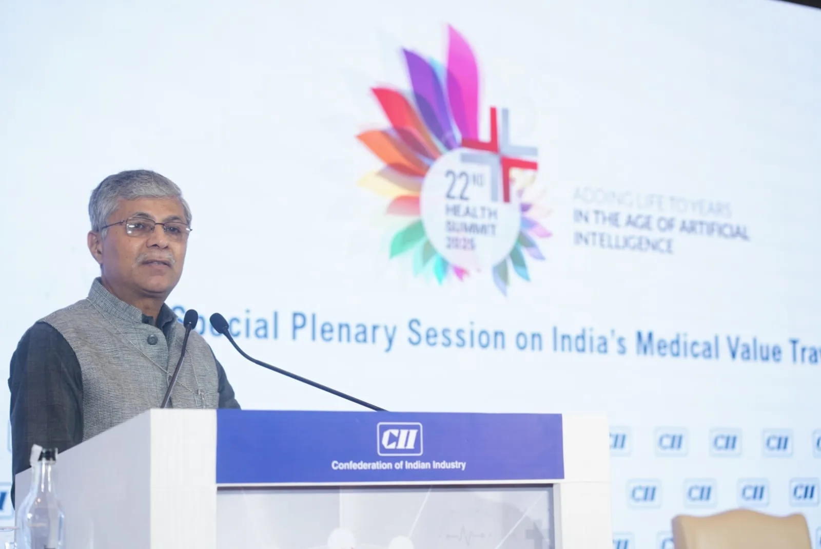 A man in a grey vest and dark shirt speaks at a podium with a microphone, in front of a large screen displaying "22nd Health Summit 2023" and "Confederation of Indian Industry".