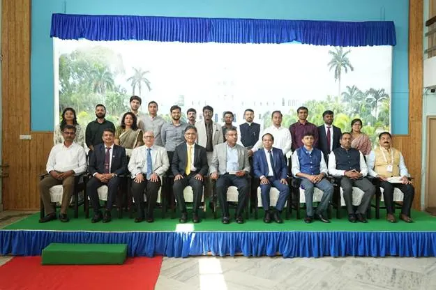 A large group of men and women, mostly men, are posing for a group photo indoors. They are dressed in business casual attire and are seated in two rows, with some standing in the back. A blue curtain is draped behind them, and a white backdrop with trees and a building is visible.