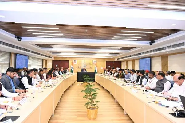 A large conference room with two long tables facing each other, filled with people in formal attire, actively participating in a meeting, with flags and screens visible in the background.