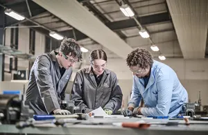 Three young students, two male and one female, wearing protective eyewear and work overalls, are focused on a project at a workbench in a workshop.