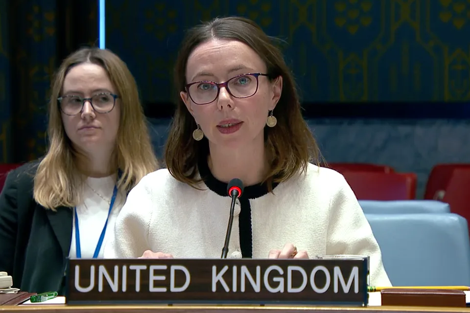 A woman with brown hair and glasses speaking at a podium with "UNITED KINGDOM" written on it. Another woman with blonde hair and glasses is seated behind her.