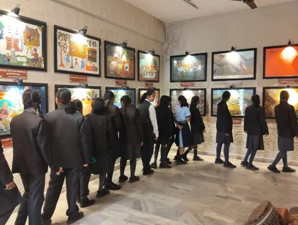 Students in dark school uniforms stand in a line, looking at artwork displayed on a wall in a brightly lit art gallery or exhibition hall. The walls are lined with framed paintings and spotlights illuminate the artwork.