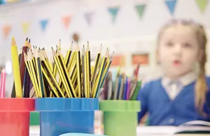 A close-up shot of yellow and black pencils in a blue cup, with other colored pencils in red and green cups, and a blurred young girl in a blue uniform sitting at a desk in the background.