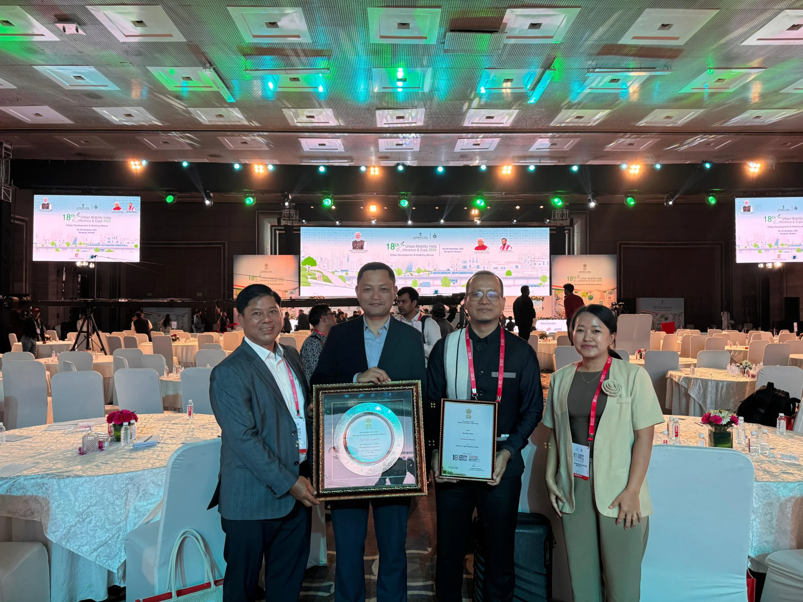 Four individuals, two men and two women, stand in a large event hall, holding awards and certificates. The man on the far left wears a grey suit and holds a framed award. The man next to him, wearing a dark suit, holds a large circular award plaque. The man on the right, wearing a dark shirt with a patterned sash, holds a framed certificate. The woman on the far right wears a light brown top and smiles at the camera. In the background, there are numerous round tables with white tablecloths and chairs, and a stage with large screens displaying graphics. The lighting in the hall is bright, with spotlights on the stage.