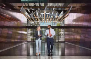  A man and a woman in business attire walk together, conversing in front of a modern building entrance.