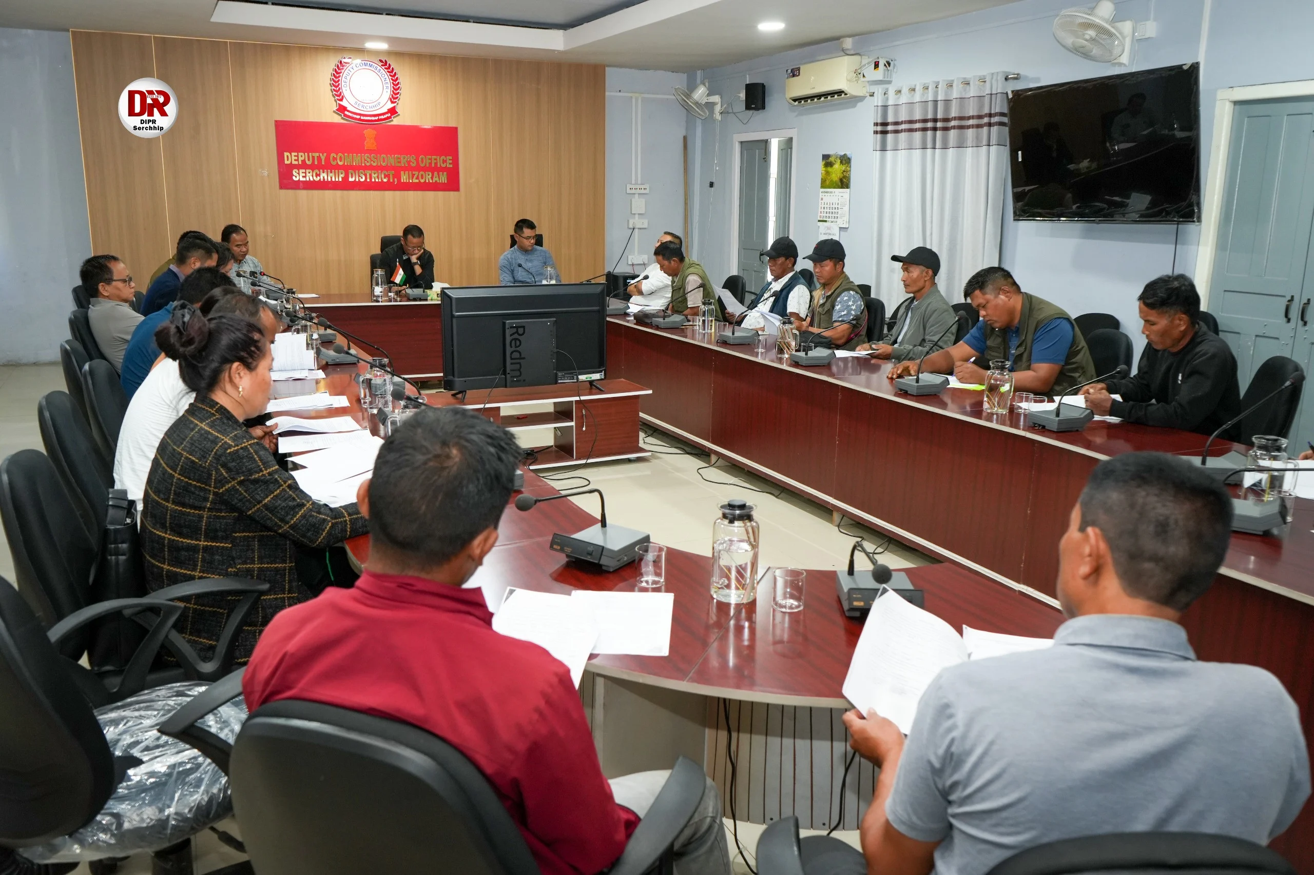 A group of people, primarily men, seated around a large U-shaped conference table in a meeting room. A red sign on the wall reads "DEPUTY COMMISSIONER'S OFFICE SERCHHIP DISTRICT, MIZORAM." Some attendees are taking notes or looking at documents.