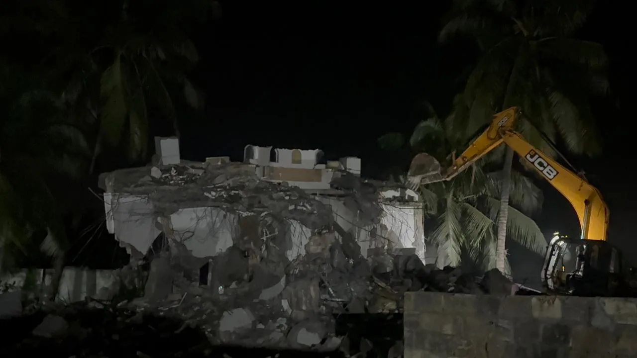 A nighttime scene shows a yellow JCB excavator working on the demolition of a white concrete structure, with palm trees visible in the background against a dark sky.
