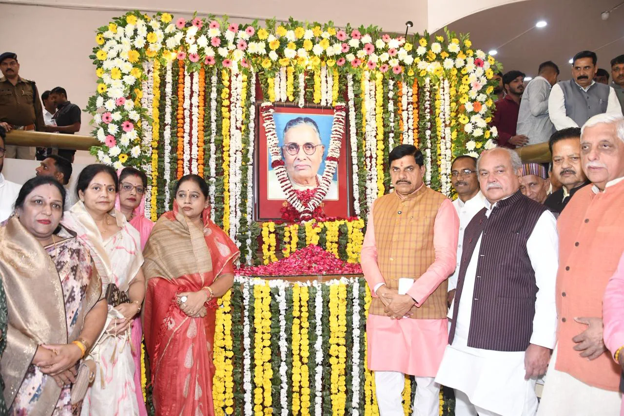 A group of people, including men and women, standing in front of a garlanded portrait of a man, likely at a commemorative event. The portrait is adorned with red and yellow flowers, and there are additional garlands creating a festive backdrop. The people in the foreground are dressed in traditional Indian attire.