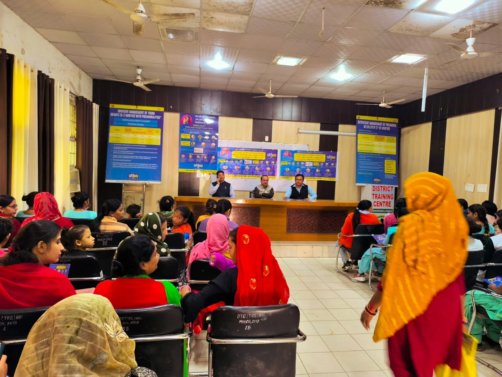 A group of women attending a training session or seminar indoors, with three men presenting from a raised platform in front of several informational posters.