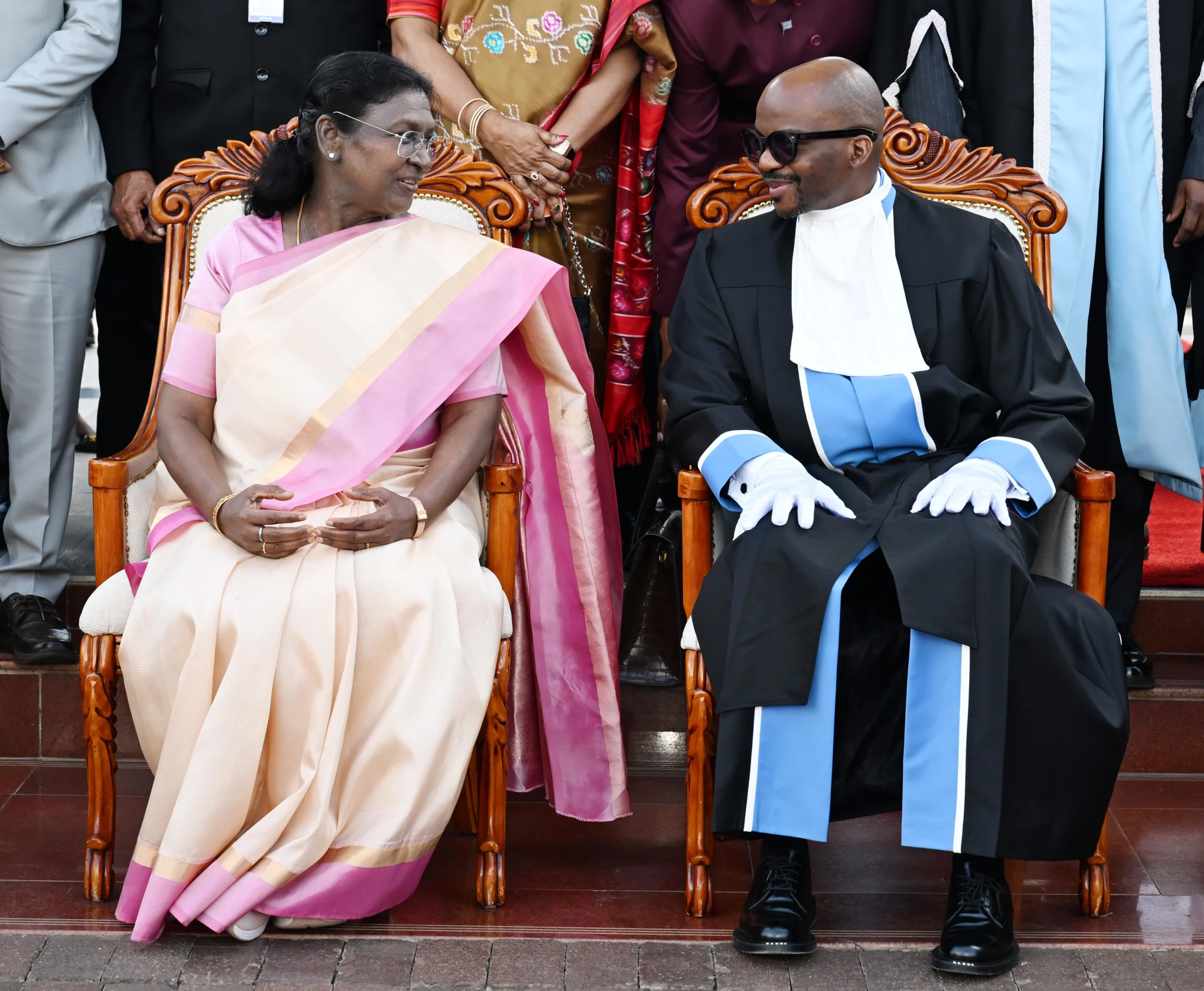 Indian President Droupadi Murmu, dressed in a traditional sari, converses with a formally robed man wearing sunglasses, both seated in ornate chairs.