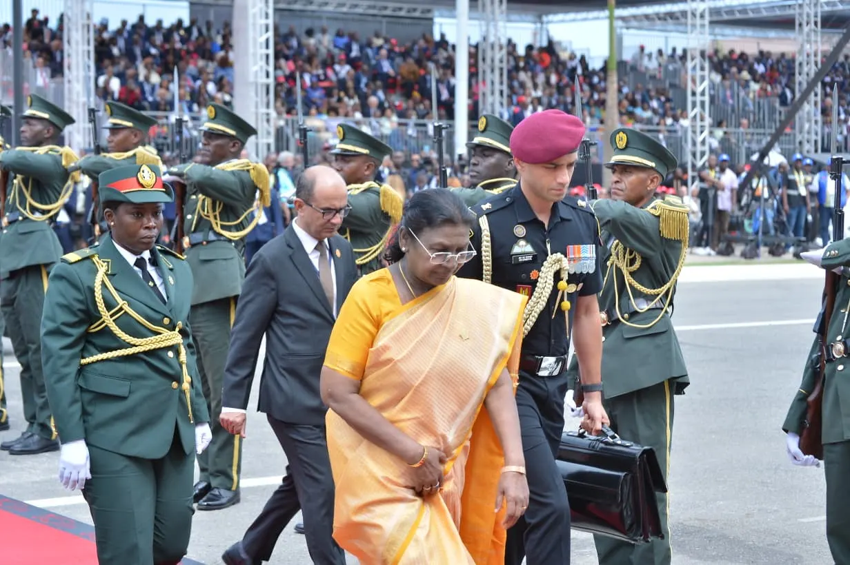 Droupadi Murmu, the President of India, walks in a procession surrounded by military personnel and other officials during a parade. She is wearing a yellow and orange saree.