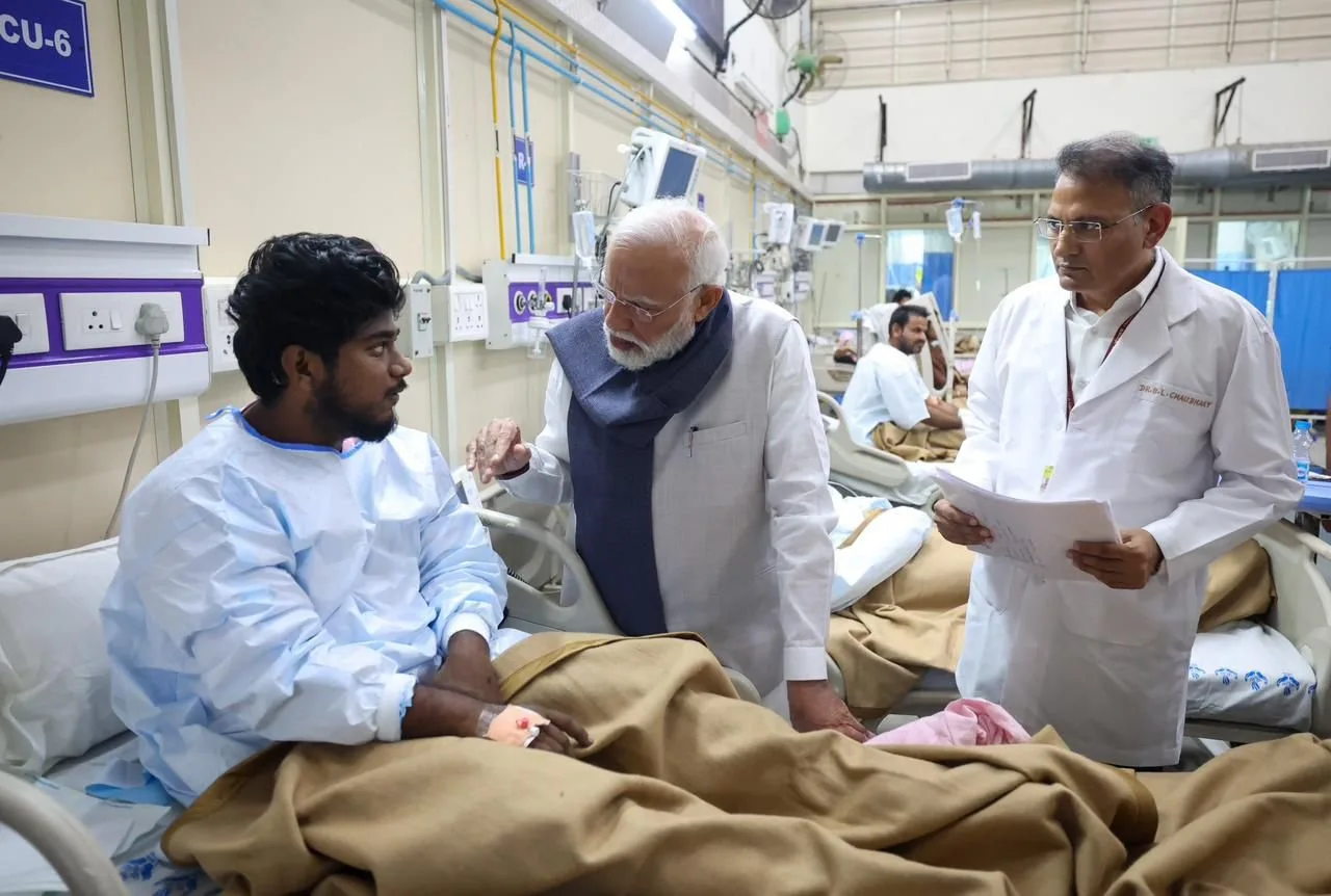 Indian Prime Minister Narendra Modi, wearing a white kurta and a dark blue shawl, speaks with a young male patient in a hospital bed. A doctor in a white coat stands next to Modi, holding documents and looking at the patient. Other patients are visible in beds in the background of the hospital ward.