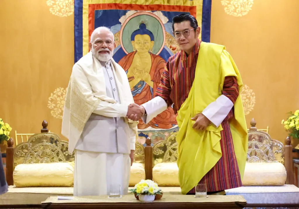 Indian Prime Minister Narendra Modi shaking hands with Bhutanese Prime Minister Tshering Tobgay, both are dressed in traditional attire, against a backdrop of traditional artwork.