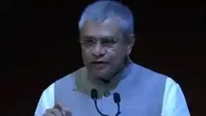 A man with short grey hair and glasses, wearing a light-colored vest over a white shirt, speaking into two microphones with a dark background.