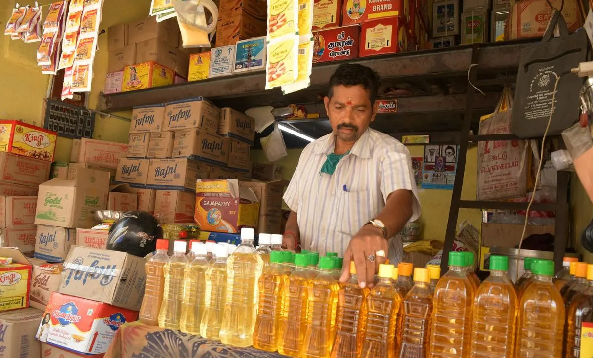 A shopkeeper stands behind a counter filled with bottles of cooking oil, with shelves of packaged goods and boxes in the background.
