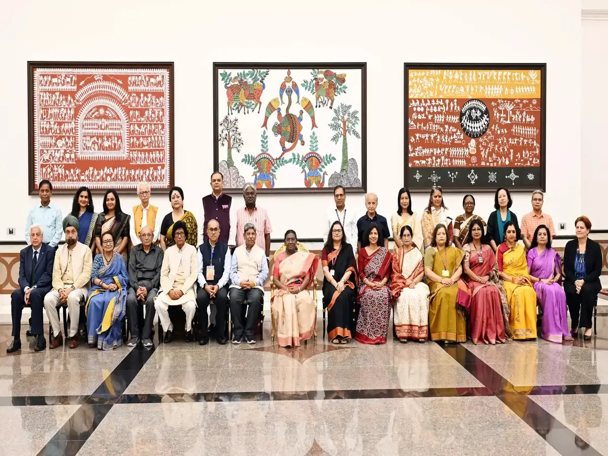 A large group of men and women, possibly delegates or officials, are seated and standing in a formal indoor setting with three large framed artworks on the wall behind them. The artwork features traditional Indian tribal art styles with intricate figures and patterns in red, black, and yellow hues.