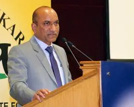  A man in a light grey suit and purple tie speaking at a wooden podium with a yellow and green banner in the background.