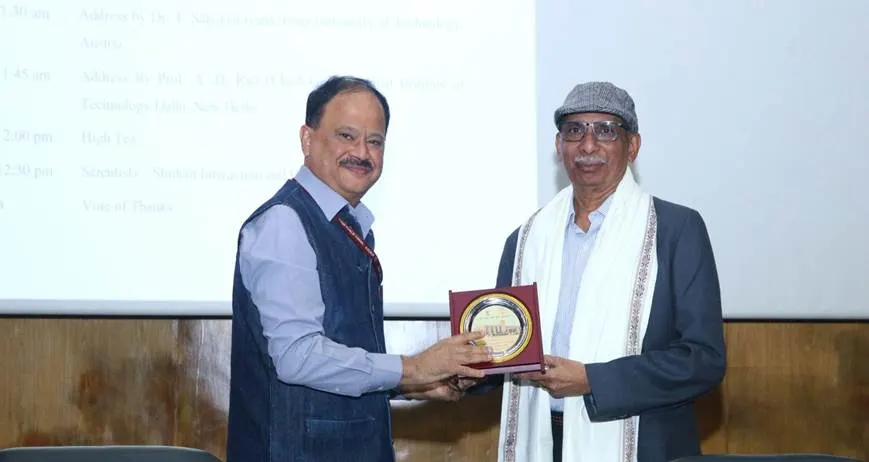 Two men, one in a blue shirt and vest, and the other in a suit jacket, white scarf, and hat, exchanging a framed plaque at an event.