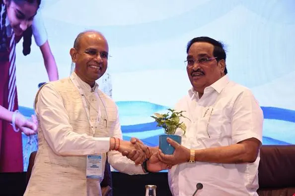 Two men, one in a white kurta and the other in a white shirt, shake hands. The man in the white shirt is presenting a small potted plant to the man in the white kurta. Both men are smiling.