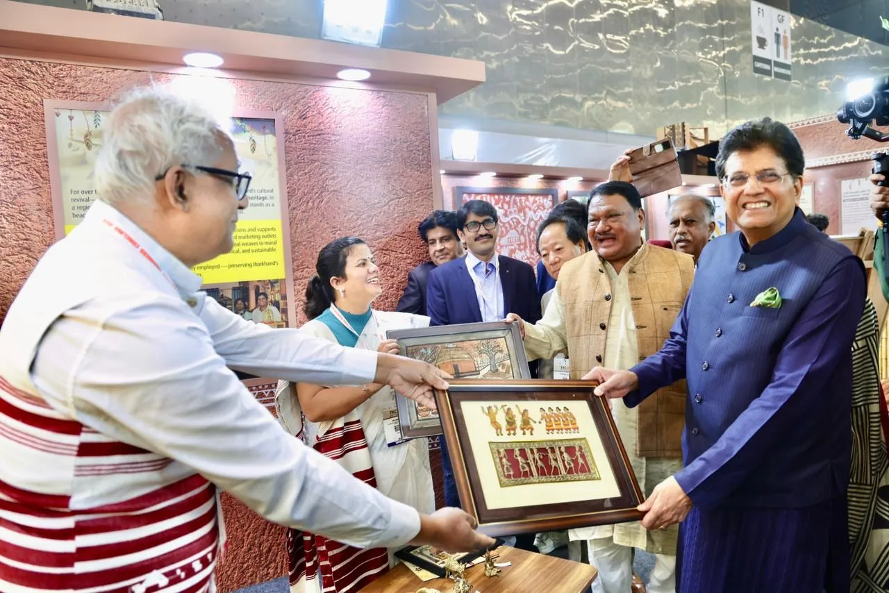 Two men exchanging framed artworks, while others observe in a well-lit indoor setting at an event.