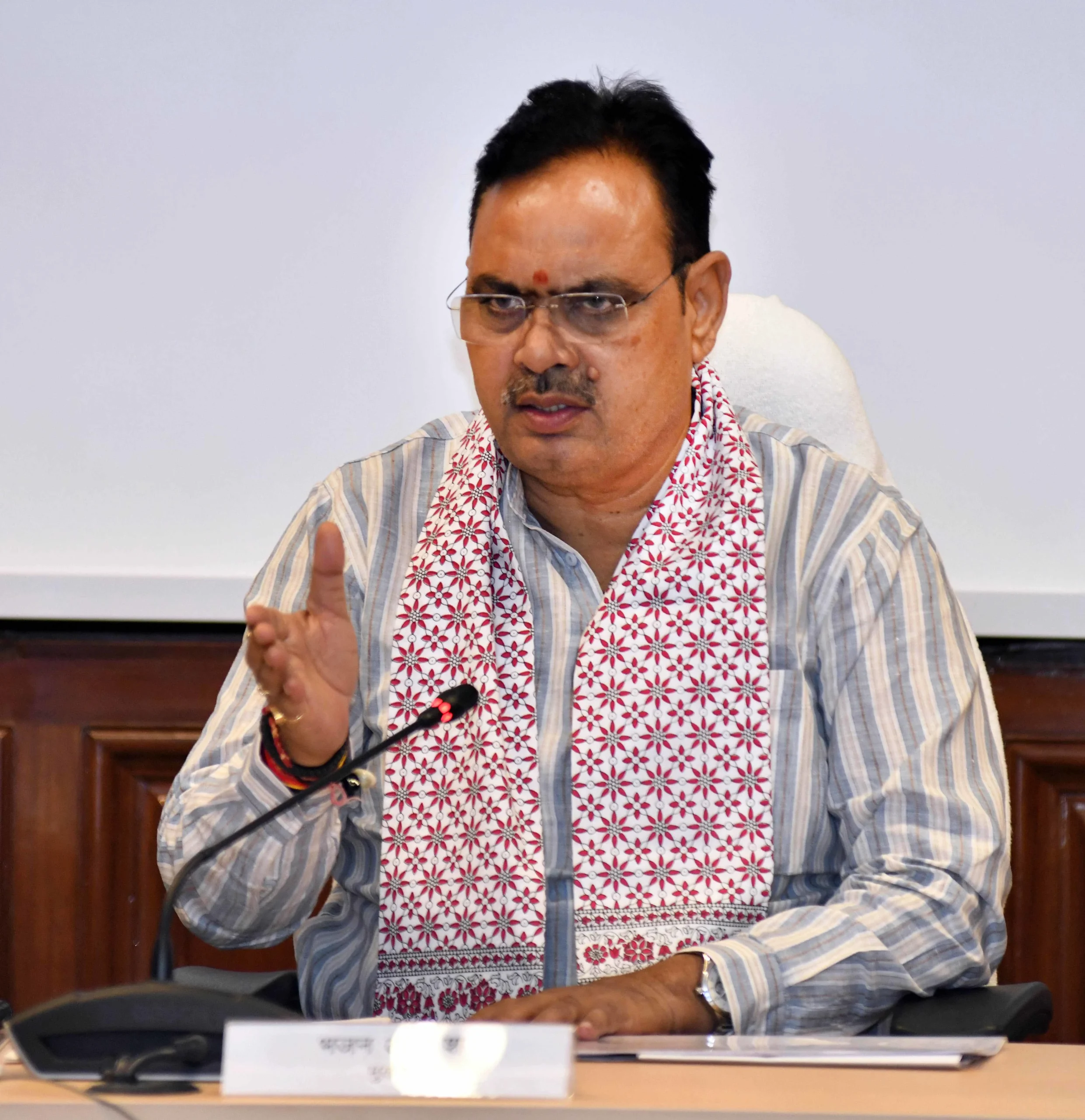 A man wearing glasses and a striped shirt with a patterned scarf around his neck is speaking at a table with a microphone in front of him. He is gesturing with his right hand.