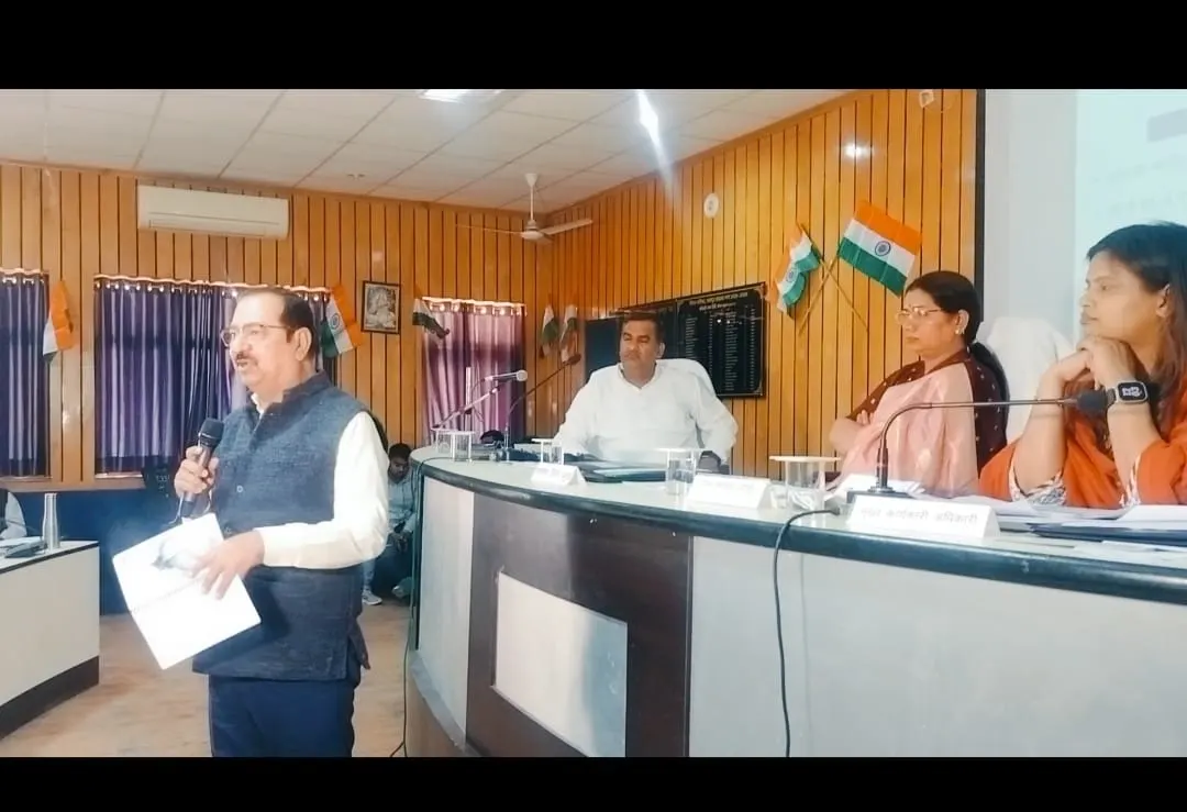 A man in a blue vest speaks into a microphone while holding papers, addressing a seated audience at a formal indoor event. Three women sit at a desk in the background, observing the speaker. Indian flags are visible on the wall behind them.