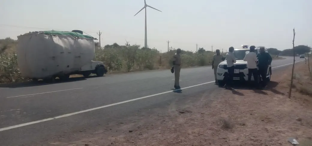 A rural road scene with a white SUV pulled over on the right shoulder, four men gathered around it, and a police officer standing in the middle of the road. On the left side of the road, a truck with a large white, covered load is parked. In the background, there are arid bushes and a large wind turbine under a clear sky.