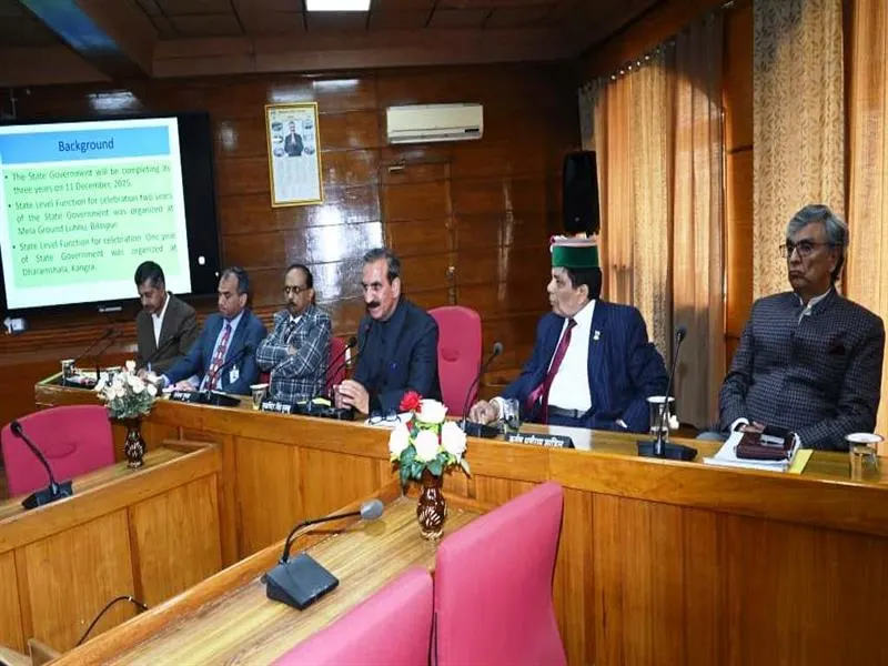 A group of five men in formal attire sitting at a long wooden table during a meeting. One man at the center is speaking into a microphone. A screen displaying "Background" and text is visible behind them.