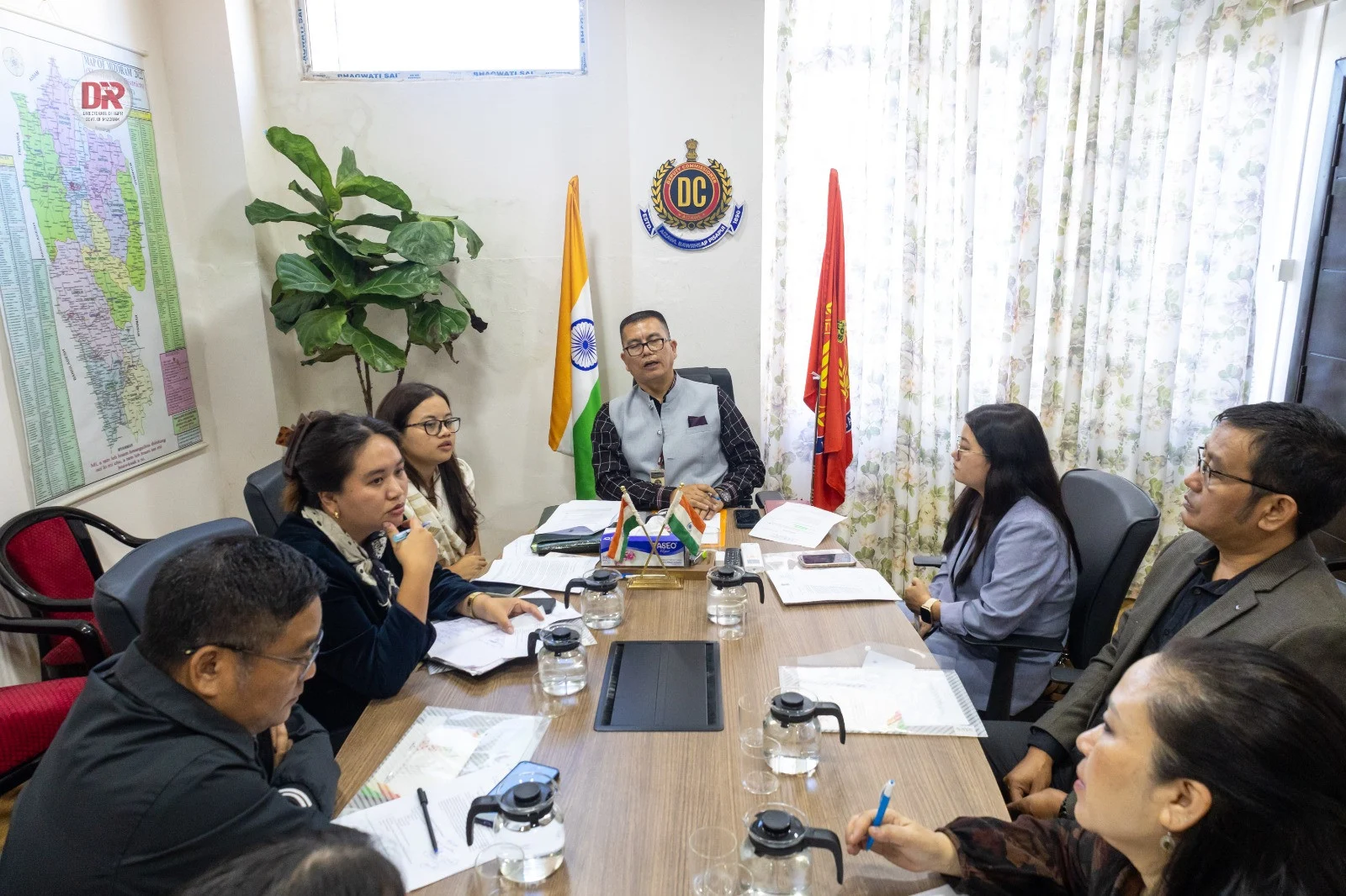 A group of officials, including men and women, attending a meeting in an office with an Indian flag and another red flag.