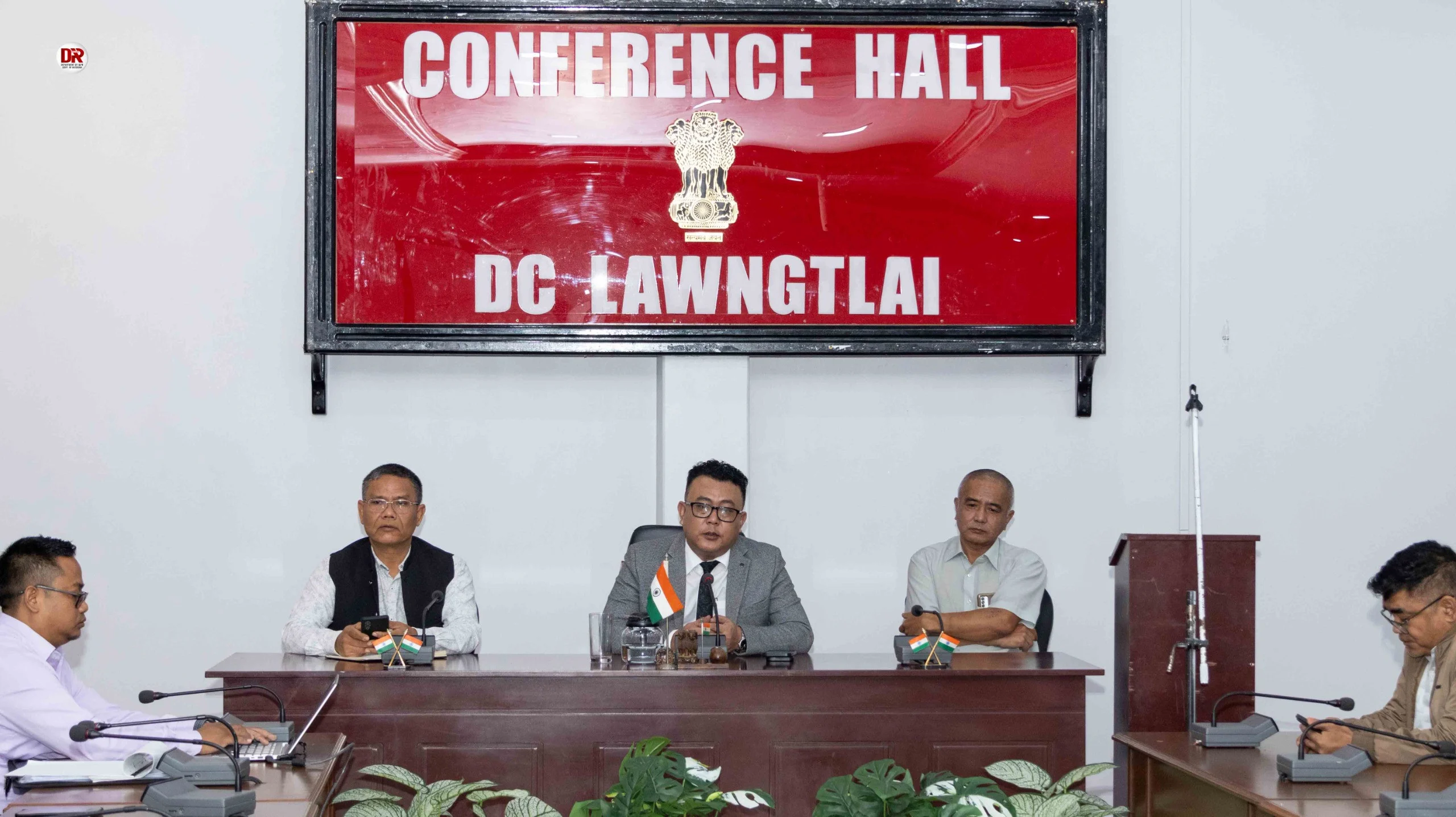 Five men are seated around a polished wooden conference table with microphones and small Indian flags. A large red sign above them reads "CONFERENCE HALL DC LAWNGTLAI".
