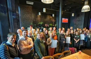 A large group of smiling professionals, mostly men and women, standing together in an indoor setting that appears to be a restaurant or event space. Some are holding papers or certificates.