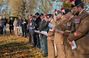 A group of people, including several soldiers in dress uniform with medals and feathered hats, standing outdoors at a memorial event, possibly a Remembrance Day ceremony. They are holding papers and looking down.