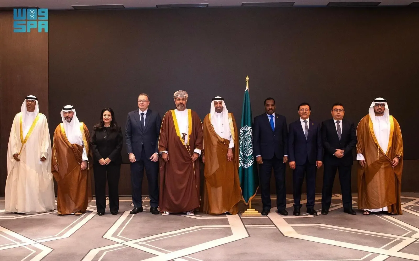 A line of eleven individuals, mostly men in suits and traditional attire, stand posing for a photo indoors, with a dark brown wall behind them and a flag on a stand to the right of the center.