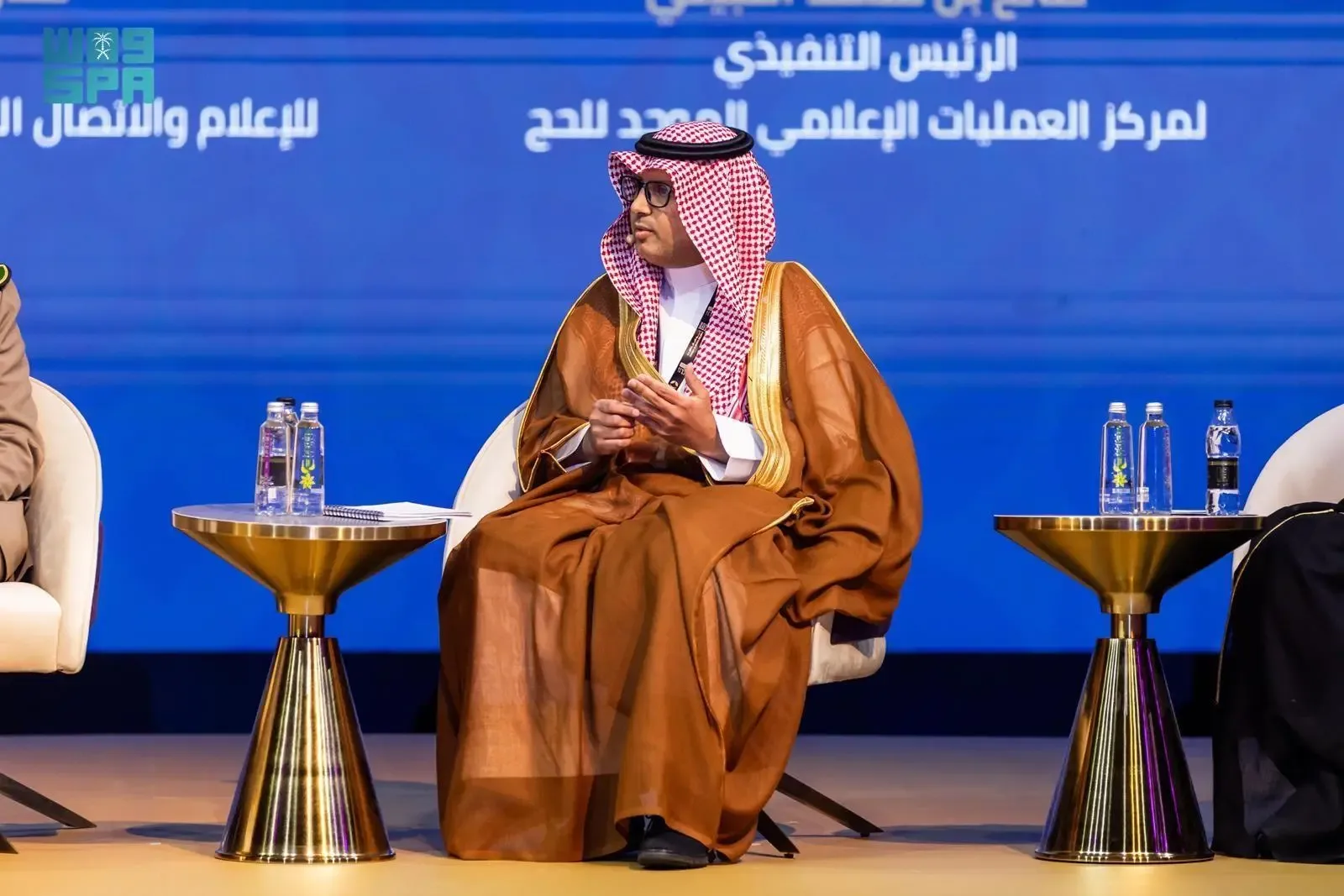 A man in traditional Saudi attire, including a thobe and ghutra, sits on a stage at a conference, looking to his left. He is wearing glasses and has a microphone in front of him. On either side of him are small golden tables with water bottles. A large blue screen with Arabic text is visible in the background.
