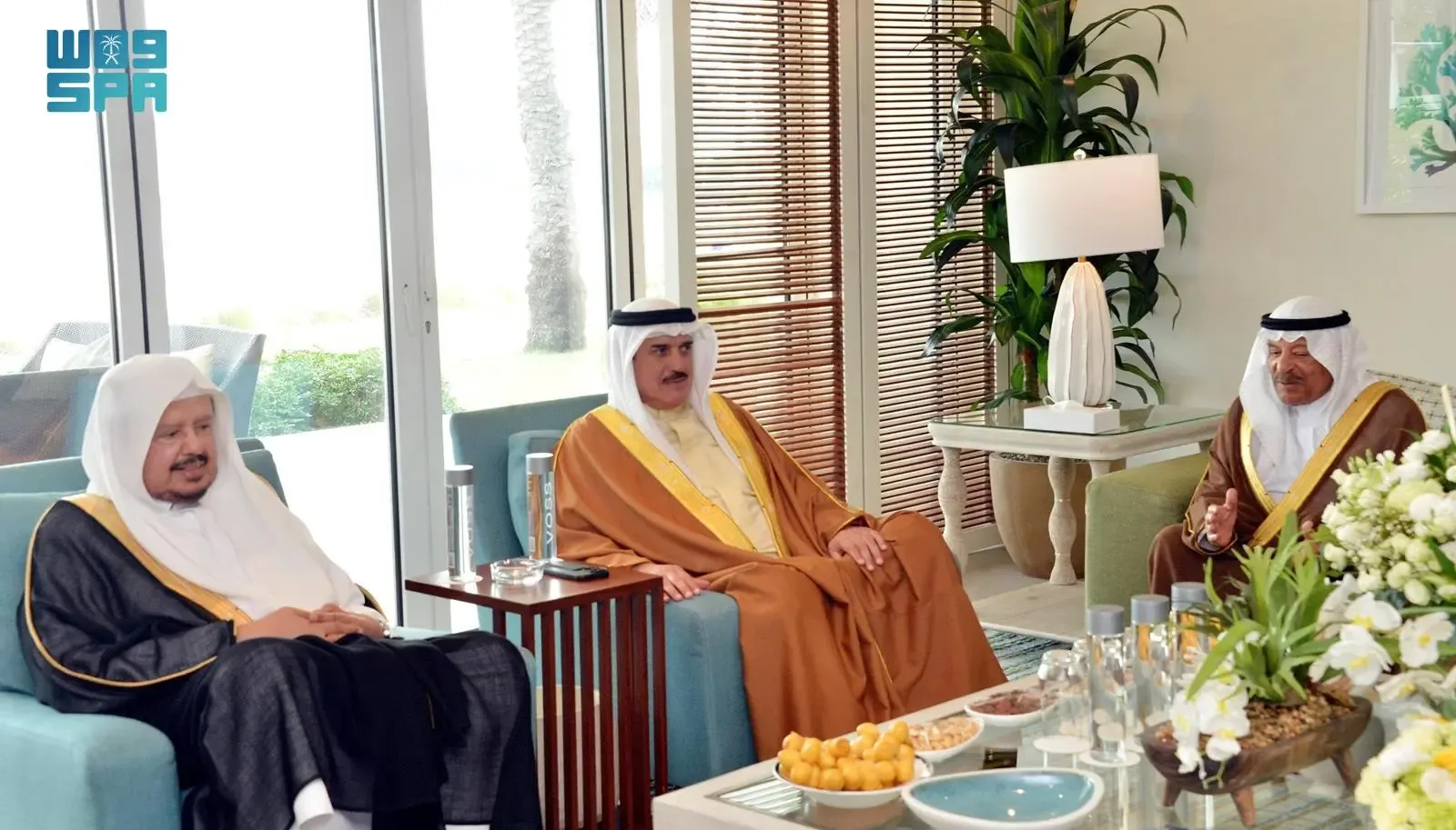 Three Saudi men in traditional thobes and ghutras seated in a modern living room, conversing.