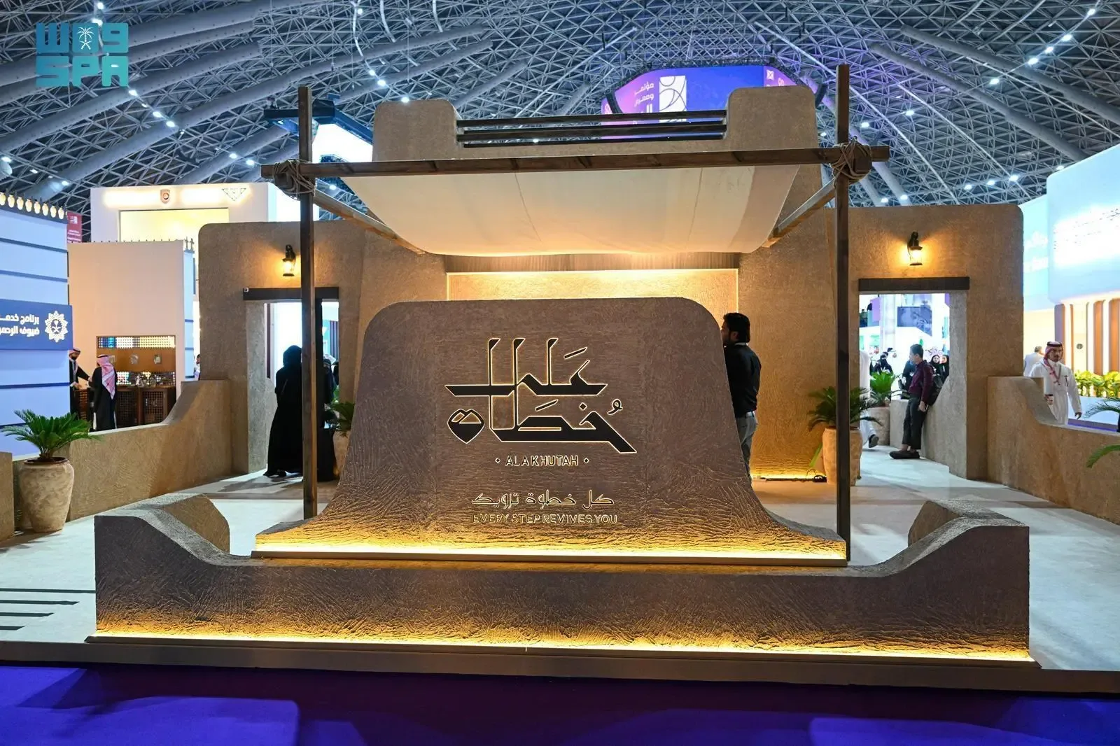A booth with a large emblem in Arabic script and "AL MAKHAWA REVIVES YOU" text, set up in an exhibition hall with people in the background.