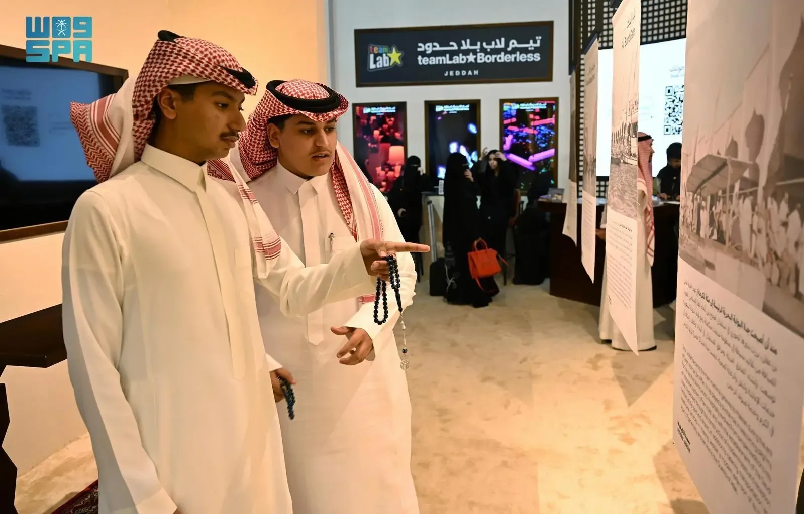 Two Saudi Arabian men in traditional thobes and ghutras looking at an exhibit, with one pointing towards a display in what appears to be a gallery or exhibition hall.