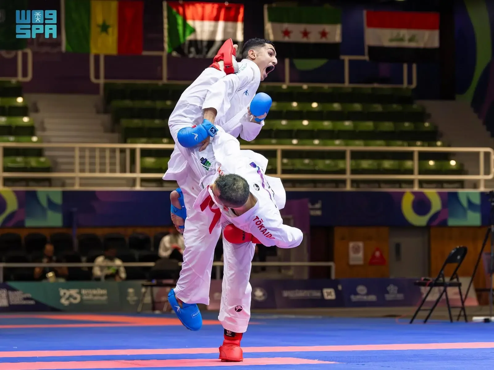 Two male karate athletes, one in white gi with blue gloves and foot protection, airborne delivering a kick to another male in white gi with red gloves and foot protection, during a competition on a blue mat in an arena with international flags in the background.