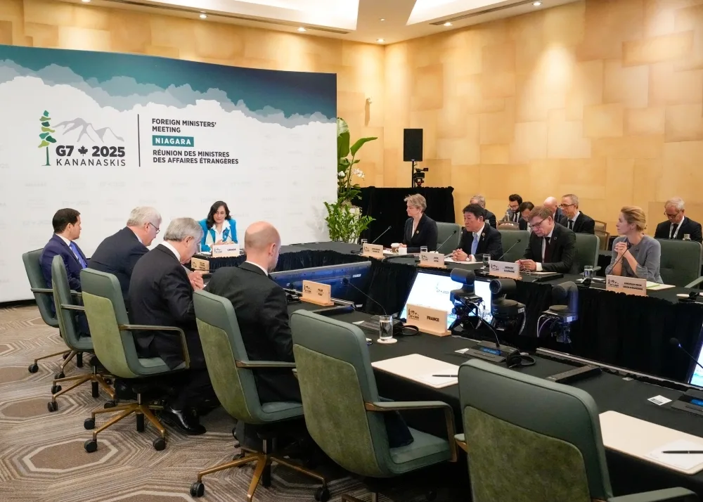 Foreign ministers and delegates attending the G7 2025 Kananaskis Foreign Ministers' Meeting in Niagara, Canada, are seated around a large conference table, engaged in discussions with nameplates and documents visible.