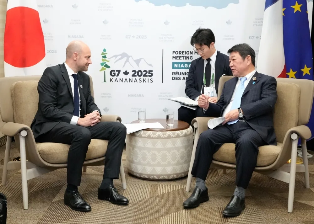 Two men in suits are seated, facing each other, in a modern conference room. A third man stands behind one of the seated figures, looking at documents. In the background, there is a sign for a "G7 2025 Kananaskis Foreign Ministers Meeting." Japanese and French flags are visible in the background.