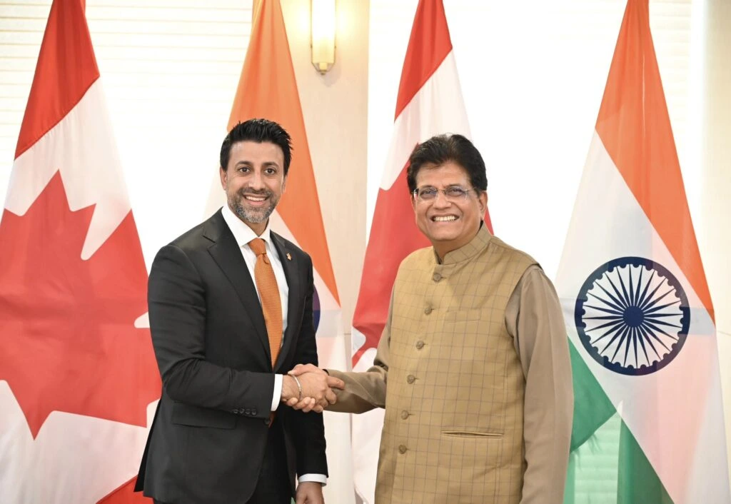 Two men in suits shaking hands in front of the Canadian and Indian flags. The man on the left is younger, with dark hair and a beard, wearing a dark suit with an orange tie and a red pin. The man on the right is older, with glasses, wearing a light brown kurta and a checkered vest.