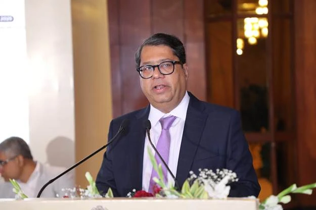 A man wearing glasses, a dark suit, and a light purple tie stands at a podium with two microphones, speaking to an audience.