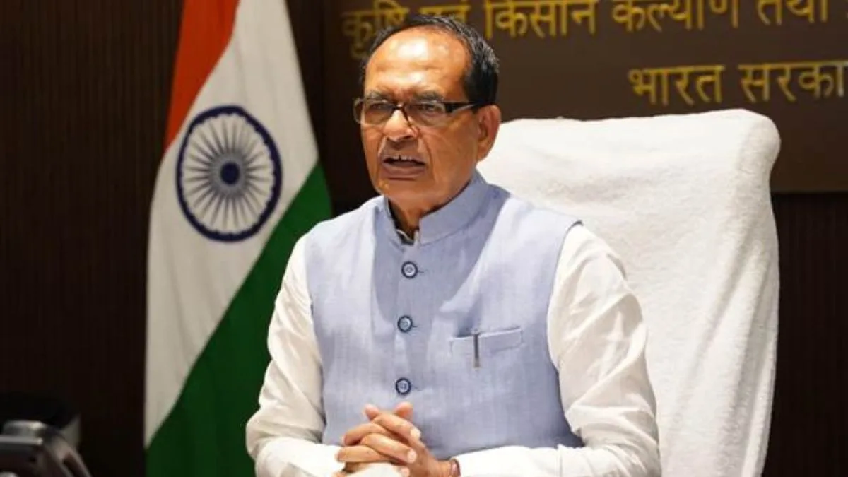 Shivraj Singh Chouhan, former Chief Minister of Madhya Pradesh, wearing a light blue vest over a white shirt, seated at a table with the Indian flag and official signage in the background.