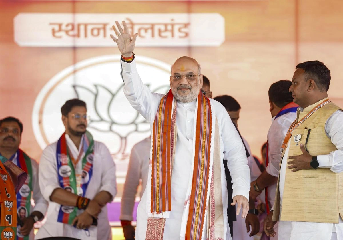 Amit Shah, an Indian politician, wearing a white kurta and an orange and white sash, waves to a crowd with his right hand raised. He is smiling and wearing glasses. Behind him is a blurred background with Hindi text and other people.