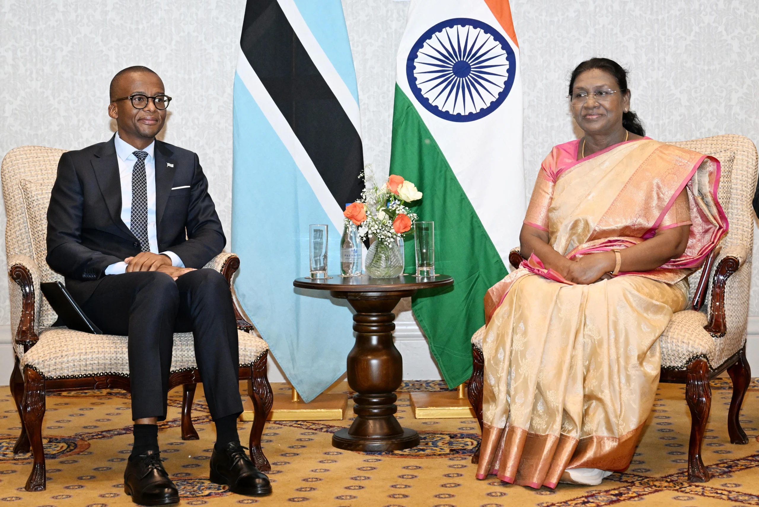 India's President Droupadi Murmu, wearing a pink and gold sari, sits opposite Botswana's Minister of Foreign Affairs, Dr. Lemogang Kwape, in a formal room with the flags of India and Botswana behind them. A small wooden table with flowers and glasses is between them.